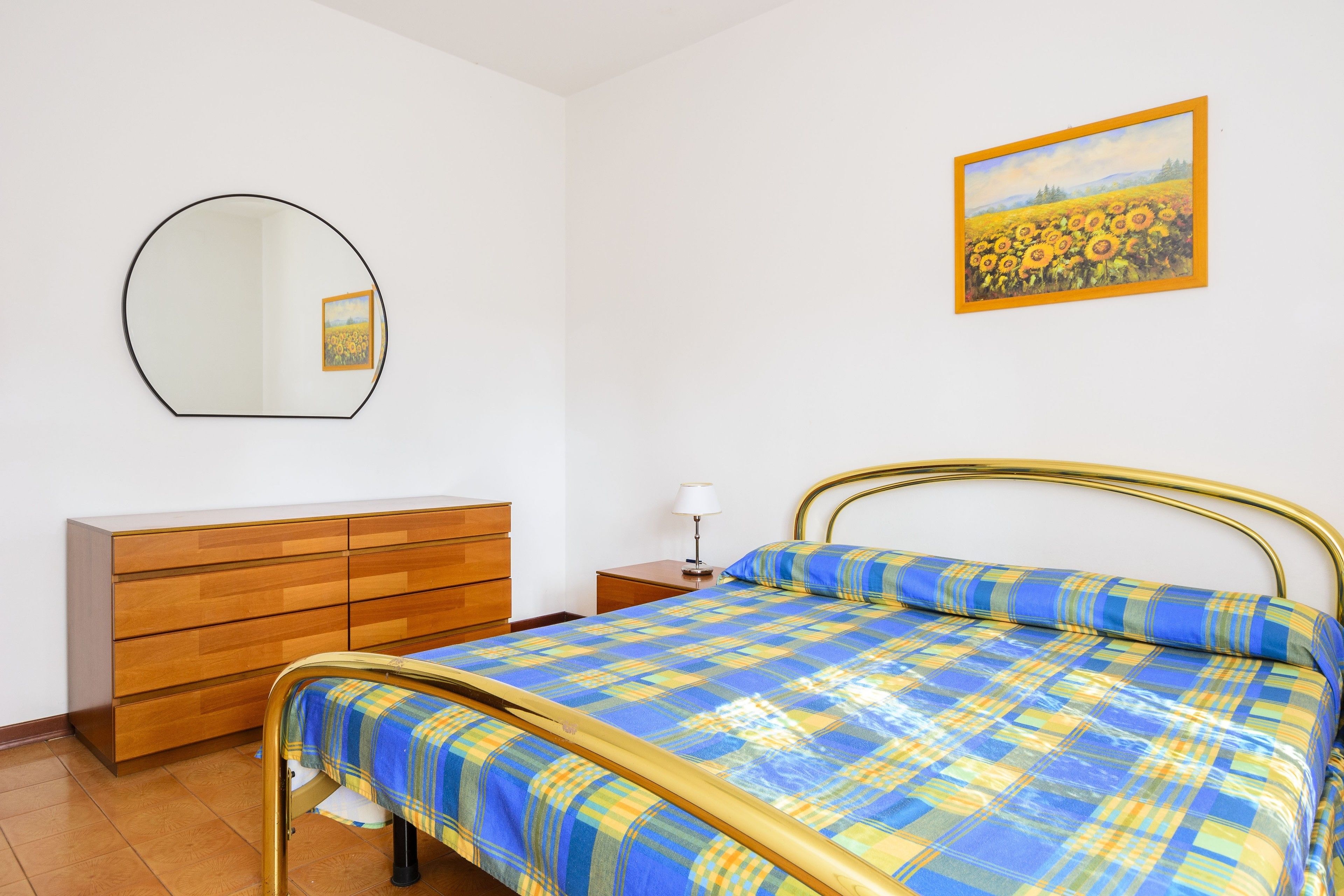 A bedroom with a bed, a dresser, a mirror, and a painting of sunflowers on the wall.