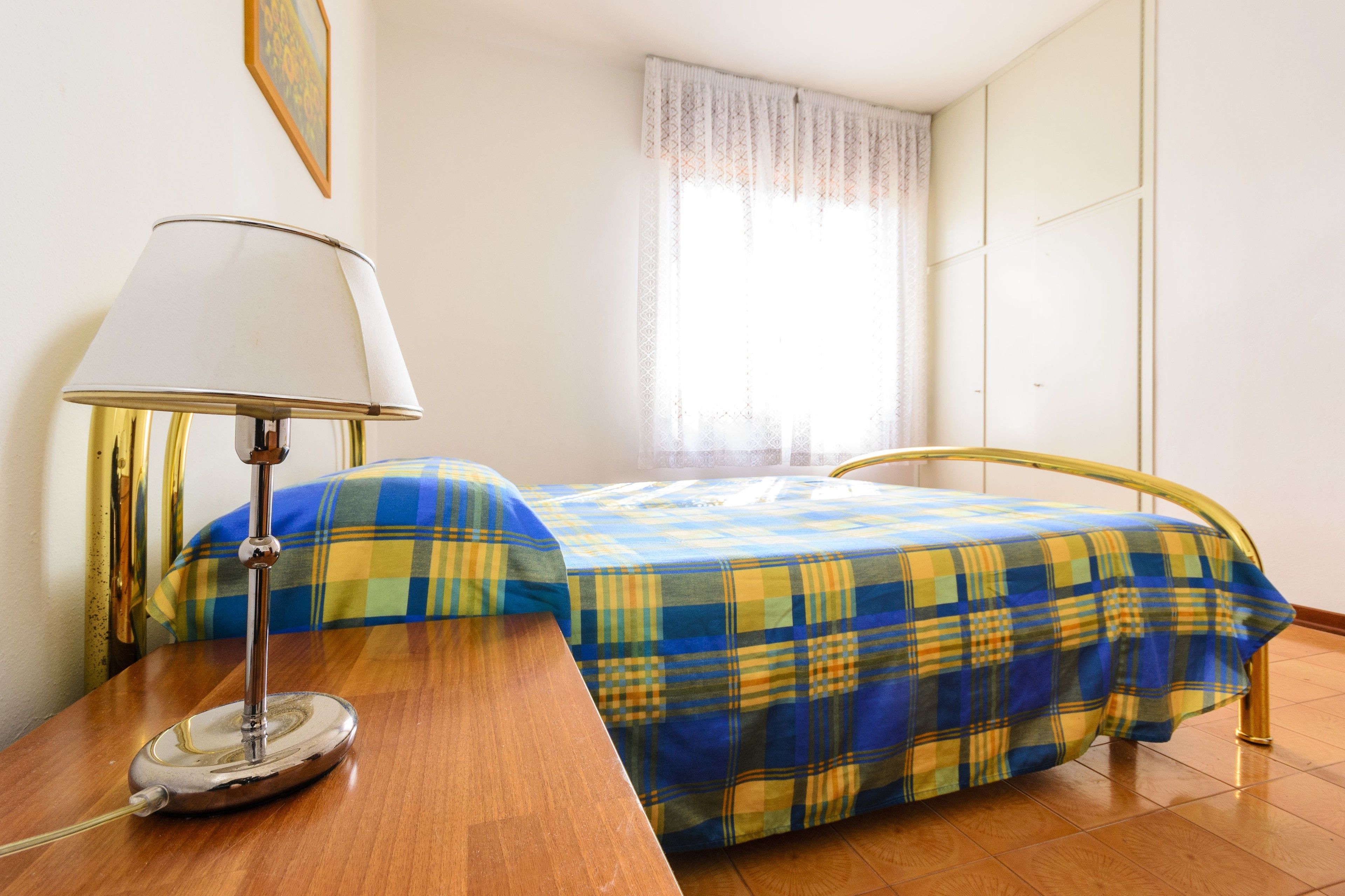 A bedroom with a bed, a bedside table with a lamp, a window with curtains, and a wall-mounted picture.