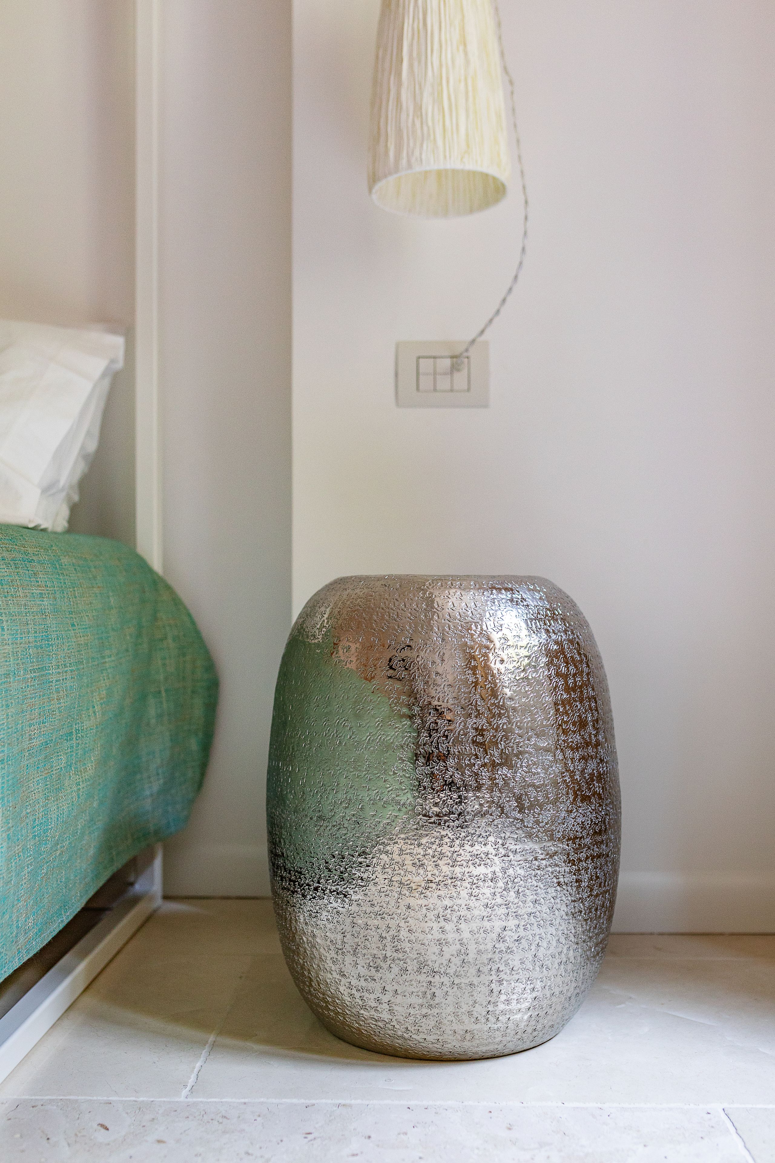 A ceramic vase on a tiled floor next to a bed with a green cover.
