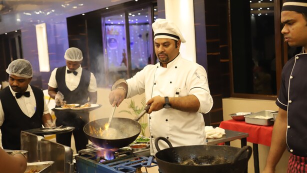 A chef cooking in a professional kitchen with stainless steel equipment and a red tablecloth. - Hotel Pooja International (Davanagere)