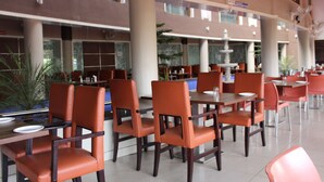 A spacious indoor dining area with multiple tables and chairs, large windows, and a fountain in the background. - Hotel Pooja International (Davanagere)