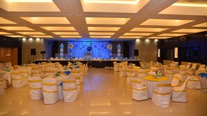 A banquet hall with round tables and chairs, a stage with blue lighting, and a ceiling with recessed lighting. - Hotel Pooja International (Davanagere)