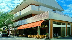 Un edificio moderno con techo plano, una cafetería con mesas al aire libre y un escaparate visible con el nombre 'ARADOR'.