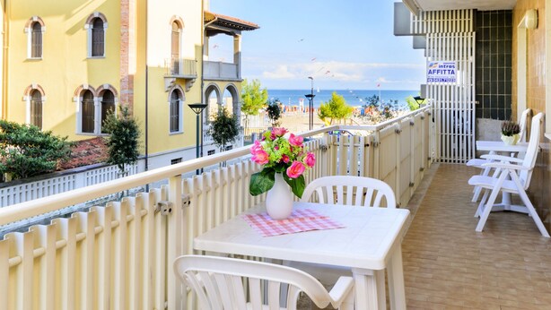 Un balcón con mesa y sillas, con vista a una playa y un edificio con ventanas arqueadas.