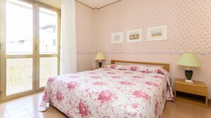 Un dormitorio con cama, mesita de noche, lámpara y ventana con vistas a un edificio.