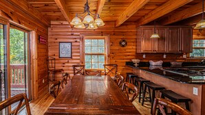 Een houten chalet van binnen, met een grote eettafel, stoelen, een keukenhoek en een groot raam.