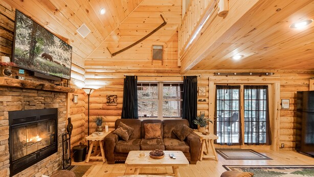 Interior acogedor de una cabaña de troncos con chimenea, televisor grande, techo de madera y zona de estar confortable.