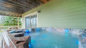 A hot tub with a wooden exterior and blue lighting, set on a porch with a green wall and a window. - Castle in the Sky 5 Bedroom House (McHenry)