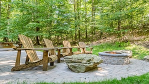 Een houten terras met Adirondack-stoelen en een vuurkorf van steen, gelegen in een bebost gebied.