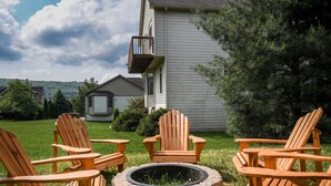 Un patio trasero con un hogar de piedra rodeado de sillas Adirondack de madera, una casa con balcón y un césped.