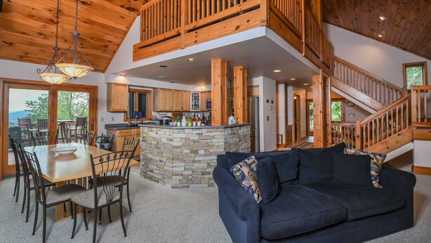A spacious living area with a high ceiling, a dining table with chairs, a dark sofa, and a stone fireplace. - Bluebird Daze 4 Bedroom House (McHenry)