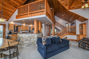 A spacious living area with a blue sofa, a dining table with chairs, a kitchen with a stone island, and a wooden staircase. - Bluebird Daze 4 Bedroom House (McHenry)