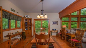 A wooden dining room with a large table, chairs, and a chandelier. There are windows with a view of trees outside.