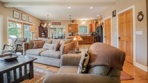 A spacious living room with a large sofa, a coffee table, and a fireplace. There is a kitchen in the background with wooden cabinets and a refrigerator. - Canadian Day 4 Bedroom House (McHenry)