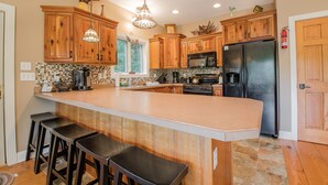 A kitchen with wooden cabinets, a large island with bar stools, a refrigerator, and a coffee maker. - Canadian Day 4 Bedroom House (McHenry)