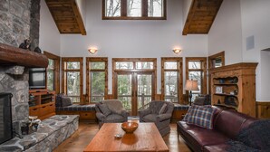 A cozy living room with a stone fireplace, wooden furniture, and large windows offering a view of the outdoors.