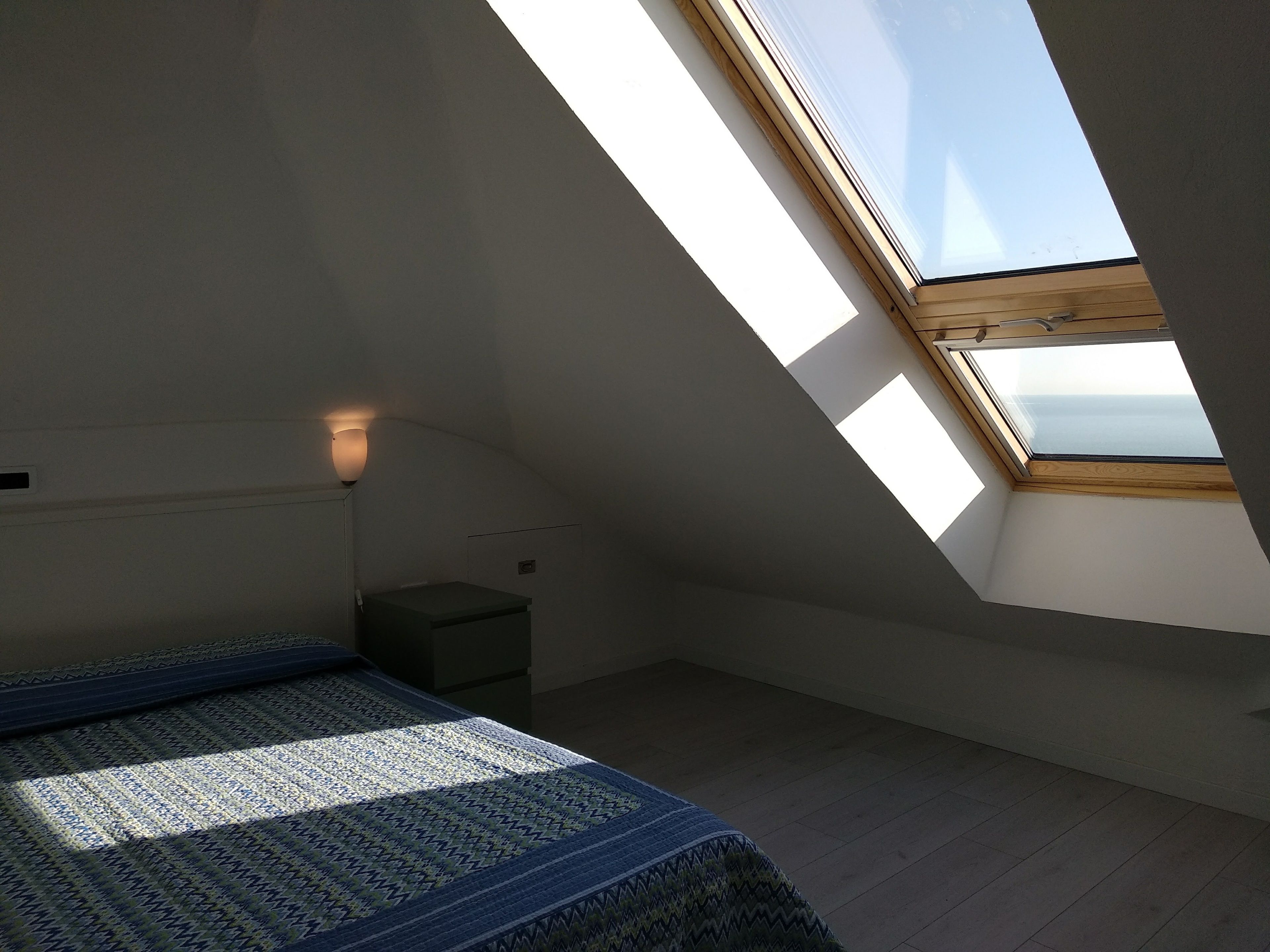 Een kleine, zwak verlichte kamer met een schuine plafond, een enkel bed, een nachtkastje en een raam met uitzicht op de lucht.