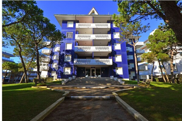 Um edifício com vários andares, uma entrada central, cercado por árvores e um gramado bem cuidado.