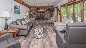 A cozy living room with a stone fireplace, comfortable seating, and large windows offering a view of the outdoors. - Lake View 3 Bedroom House (Oakland)