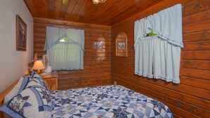 Ein Schlafzimmer in einer Holzhütte mit einem Bett, zwei mit Vorhängen versehenen Fenstern, einem Nachttisch mit Lampe und einem an der Wand befestigten Ventilator.