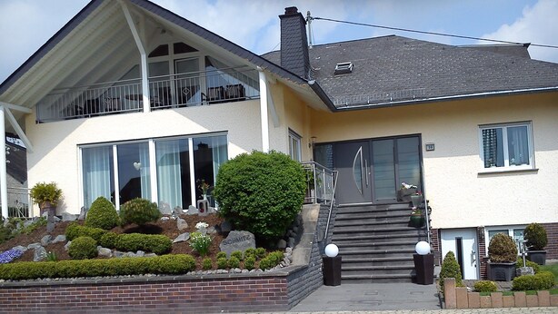 Ein zweistöckiges Haus mit Balkon, einer Treppe zum Eingang und einem gepflegten Garten mit verschiedenen Pflanzen und einer Felsformation.