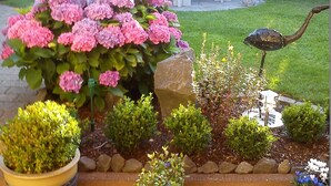 Ein Garten mit verschiedenen Kübelpflanzen, einem Kiesweg und einer kleinen Außenlounge mit Tisch und Stühlen.
