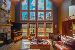 A spacious living room with a fireplace, leather sofa, and large windows offering a view of trees. - High-Skis' Villa 4 Bedroom House (McHenry)