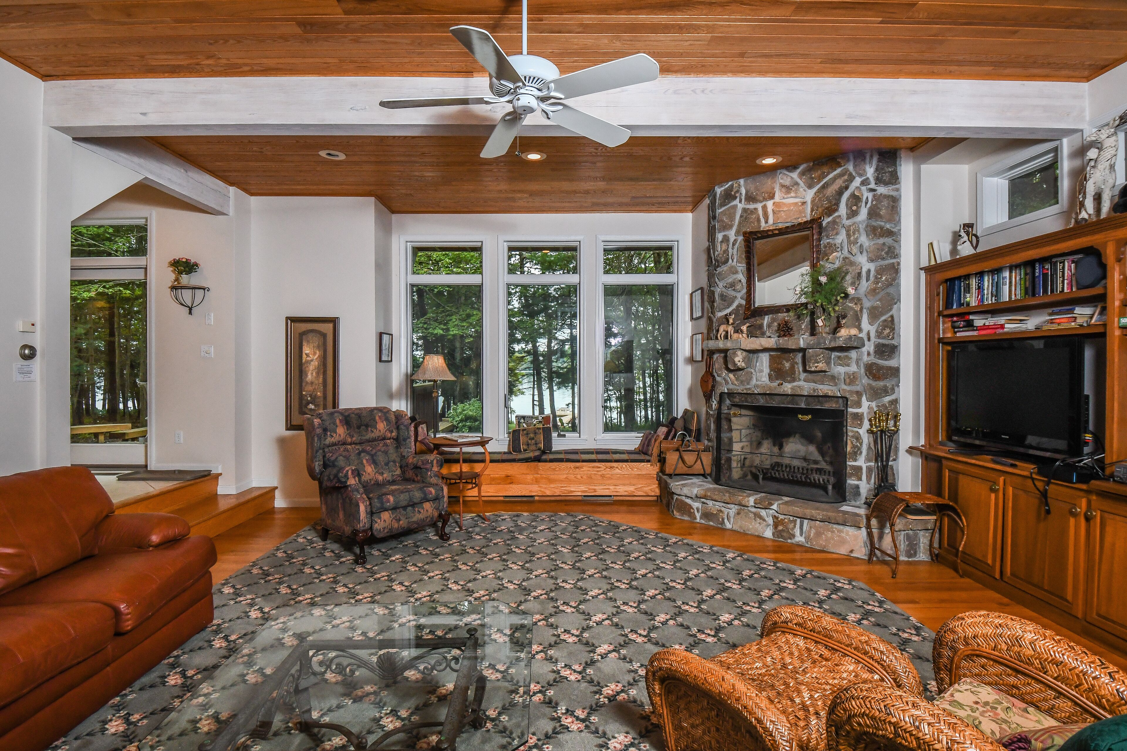 A spacious living room with a fireplace, a ceiling fan, and a large window overlooking a forest.