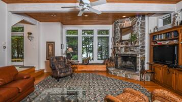 A spacious living room with a fireplace, a ceiling fan, and a large window overlooking a forest.