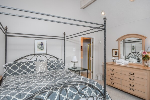 A four-poster bed with a patterned bedspread, a bedside table with a lamp, a wooden dresser with drawers, and a mirror above the dresser.