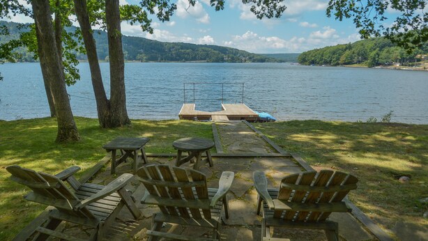 Une terrasse au bord du lac, avec des chaises et des tables en bois, offrant une vue sur un lac paisible, un quai et des collines lointaines.