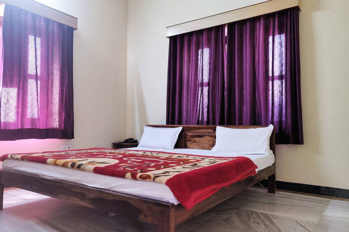 Una cama con estructura de madera, colcha estampada en rojo y beige, y almohadas blancas.