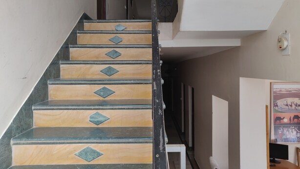 A staircase with alternating wooden and marble steps, a metal railing, and a hallway leading to a room with a bed and a desk. - Hotel Leela Nikunj (Jaisalmer)