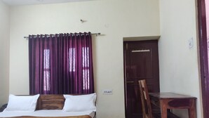 A bedroom with a bed, a wooden chair, a desk, and a door. - Hotel Leela Nikunj (Jaisalmer)