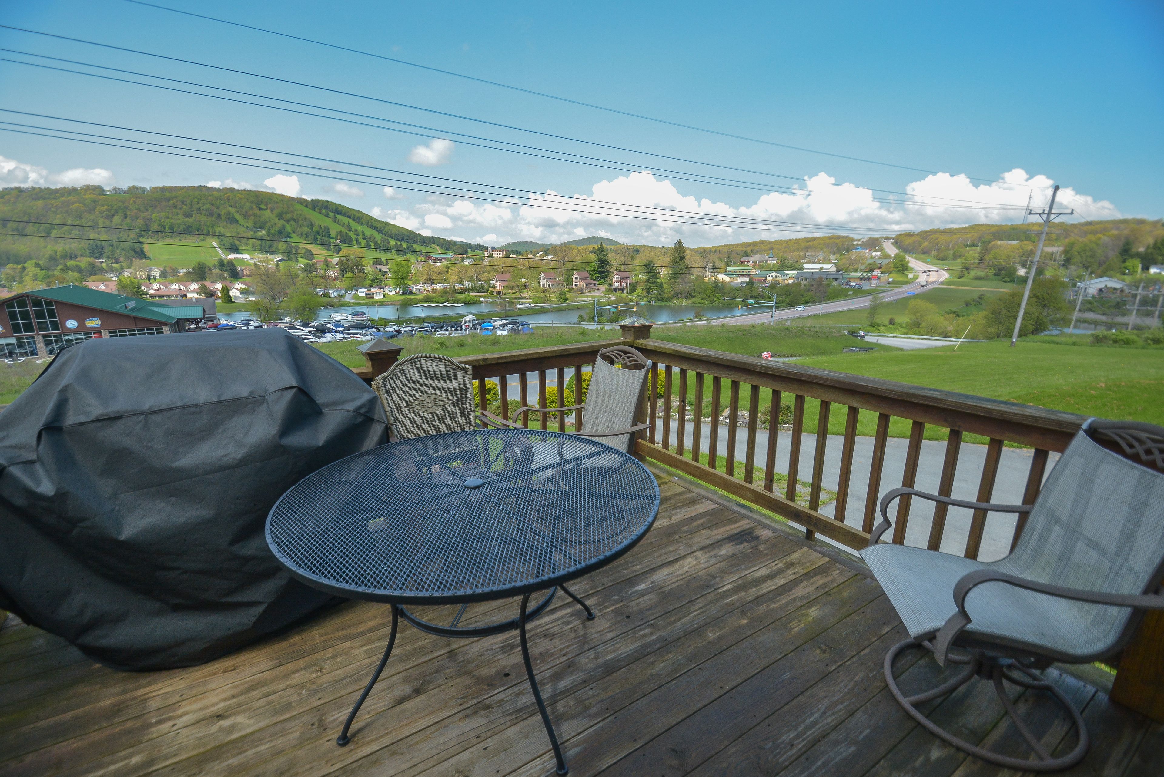 A wooden deck with a round table and two chairs, overlooking a scenic view of a lake and a town.