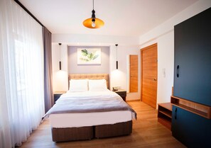 A modern hotel room with a large bed, wooden flooring, a ceiling light, a nightstand, a wall-mounted artwork, and a wardrobe.