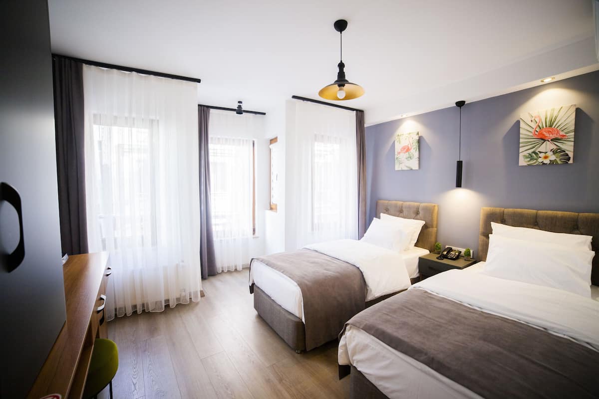 Habitación de hotel con dos camas, un escritorio de madera y una silla. Hay cortinas en las ventanas y obras de arte en las paredes.
