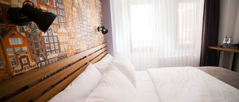 A hotel room with a neatly made bed, a headboard with a cityscape mural, a bedside lamp, and a window with sheer curtains.