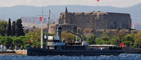 A large ship with a flag, docked near a historic stone fortress with flags.