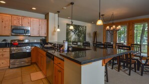 A kitchen with wooden cabinets, a stainless steel stove, and a large island with a black countertop. There are hanging pendant lights and a chandelier. - Retreat on Main Street 4 Bedroom Townhome (McHenry)