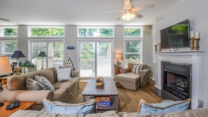 A spacious living room with a fireplace, large windows, and a ceiling fan.