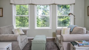 A room with two beige sofas, a small table, and a lamp. There are three windows with white curtains, and a view of trees outside.