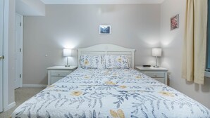A bedroom with a bed, two bedside tables, a nightstand, a picture on the wall, and a window with curtains.