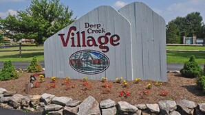 Un letrero de madera que indica Deep Creek Village, con un logotipo y un área ajardinada con flores y rocas.
