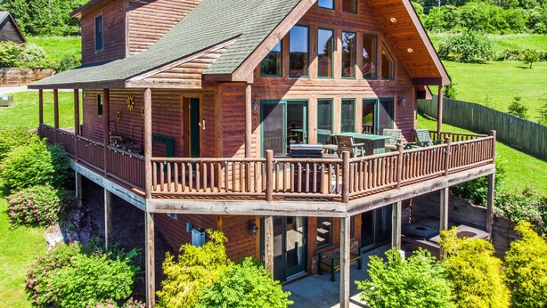 A two-story log cabin with a balcony and a deck, surrounded by lush greenery.