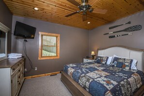 A bedroom with a wooden ceiling, a bed with a floral quilt, a nightstand with a lamp, and a TV mounted on the wall.