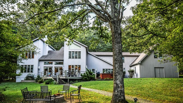 A large white house with a covered porch, a lawn with a bench, and a tree in the foreground.