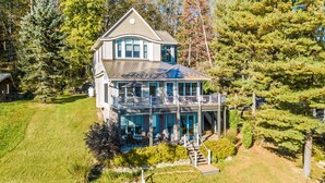 Une grande maison à deux étages, dotée d’un balcon et d’un porche couvert, entourée d’arbres et d’une pelouse.