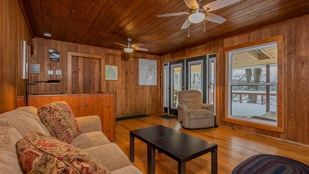 A cozy living room with wooden walls, a sofa with patterned cushions, a coffee table, and a chair. There are ceiling fans, a map on the wall, and a view through a large window. - Ski Shore 3 Bedroom House (Swanton)
