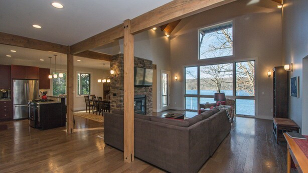 A spacious living area with a large gray sectional sofa, wooden beams, a stone fireplace, and a view of a body of water through large windows.